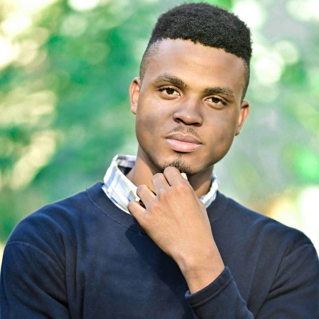 Taslan Graham is standing tall, posing with one hand under his chin, as if thinking deeply. His eyes are set on the camera, in a serious yet contemplative manner. The canopy is blurred in the background, bringing his figure in sharp focus.
