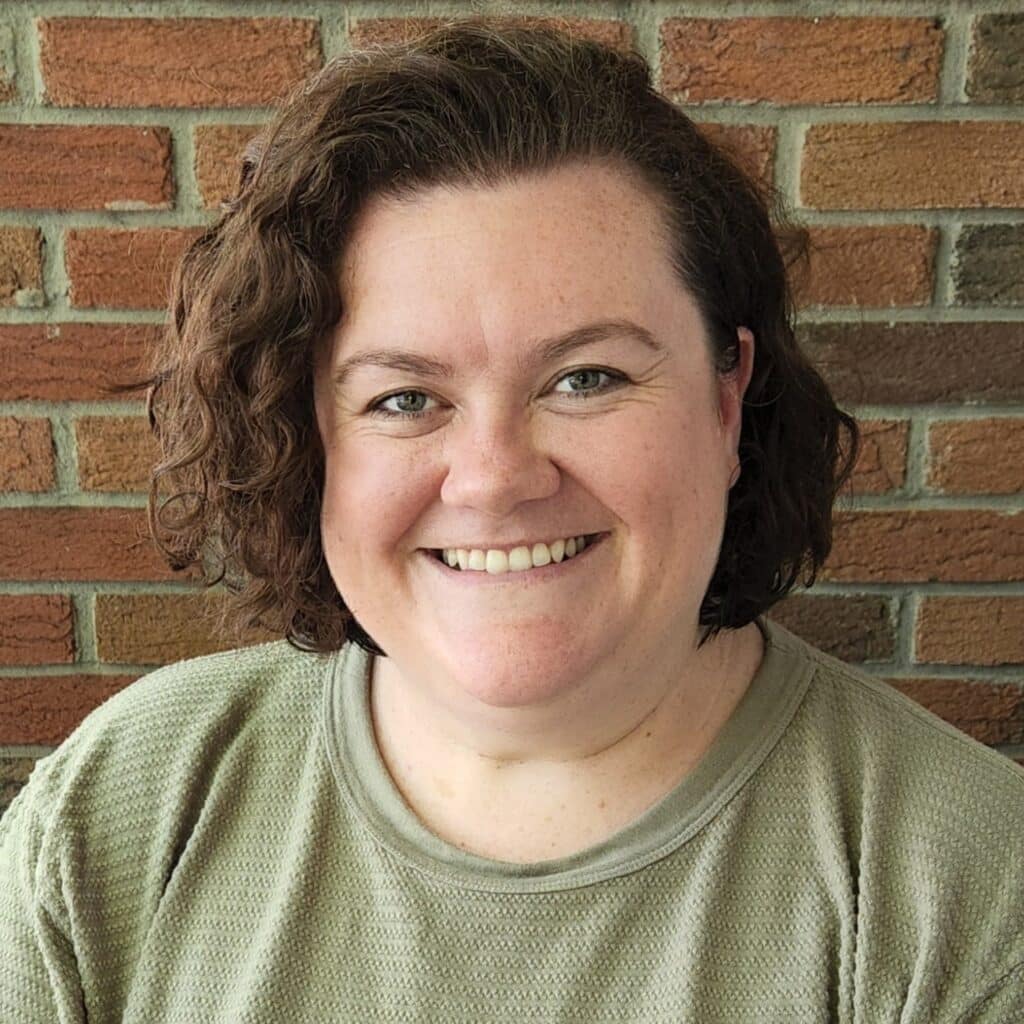 Zoe poses in front of a classic red brick wall, wearing a light green t-shirt. Her chin-length brown, curly hair frames her smiling face. The expression is one of persistence and confidence.